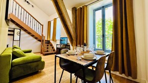 Un comedor con una mesa preparada para cuatro personas, un sofá verde, una escalera de madera y un ventanal con cortinas.