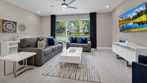 A modern living room with a grey sofa, white coffee table, and a large window with a view of greenery and mountains.