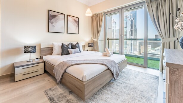 Une chambre moderne avec un grand lit, une table de chevet, un bureau et une vue sur la ville grâce à de grandes fenêtres du sol au plafond.