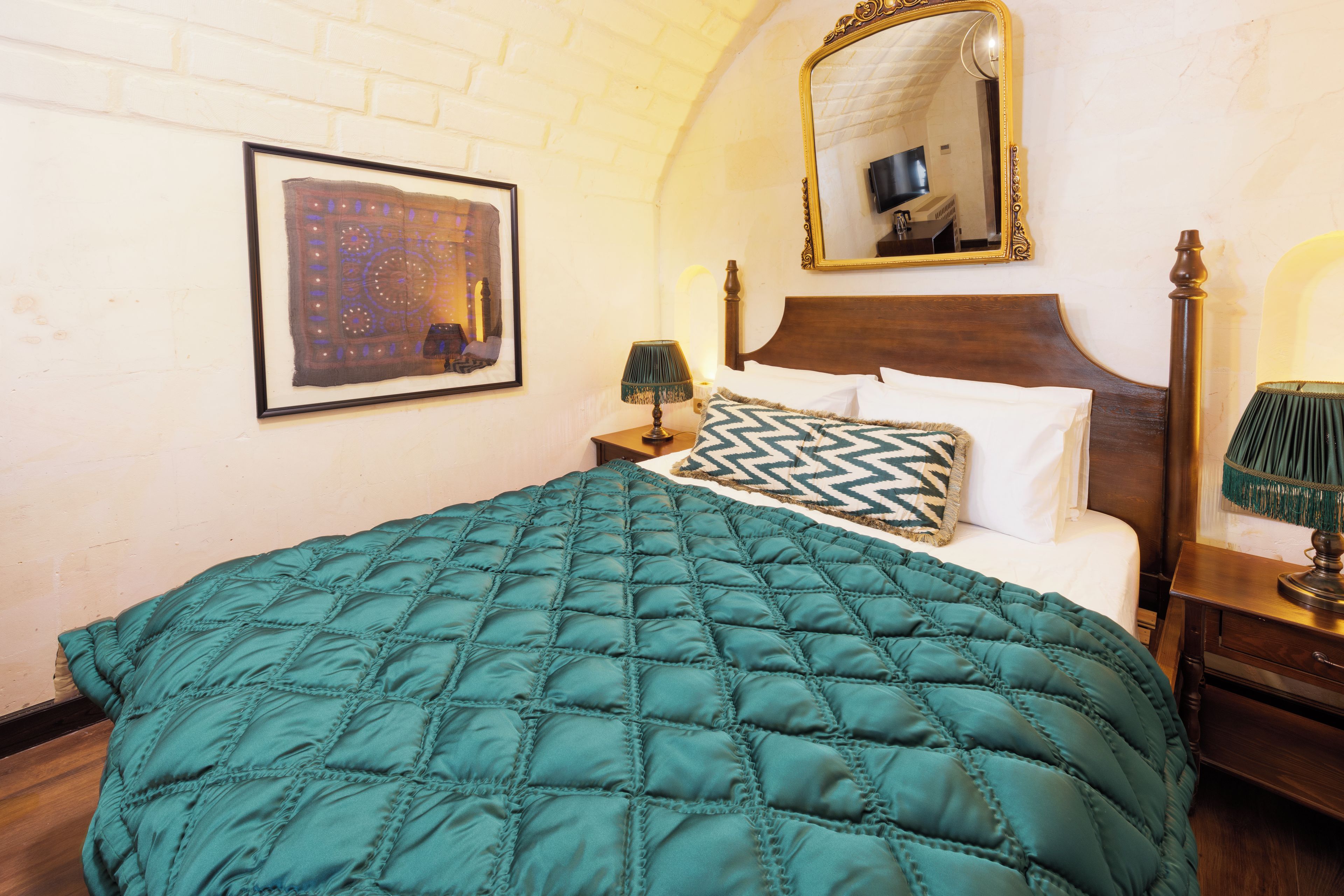 Una cama bien hecha con colcha color turquesa, cabecera de madera, una obra enmarcada en la pared y un espejo sobre la cama.