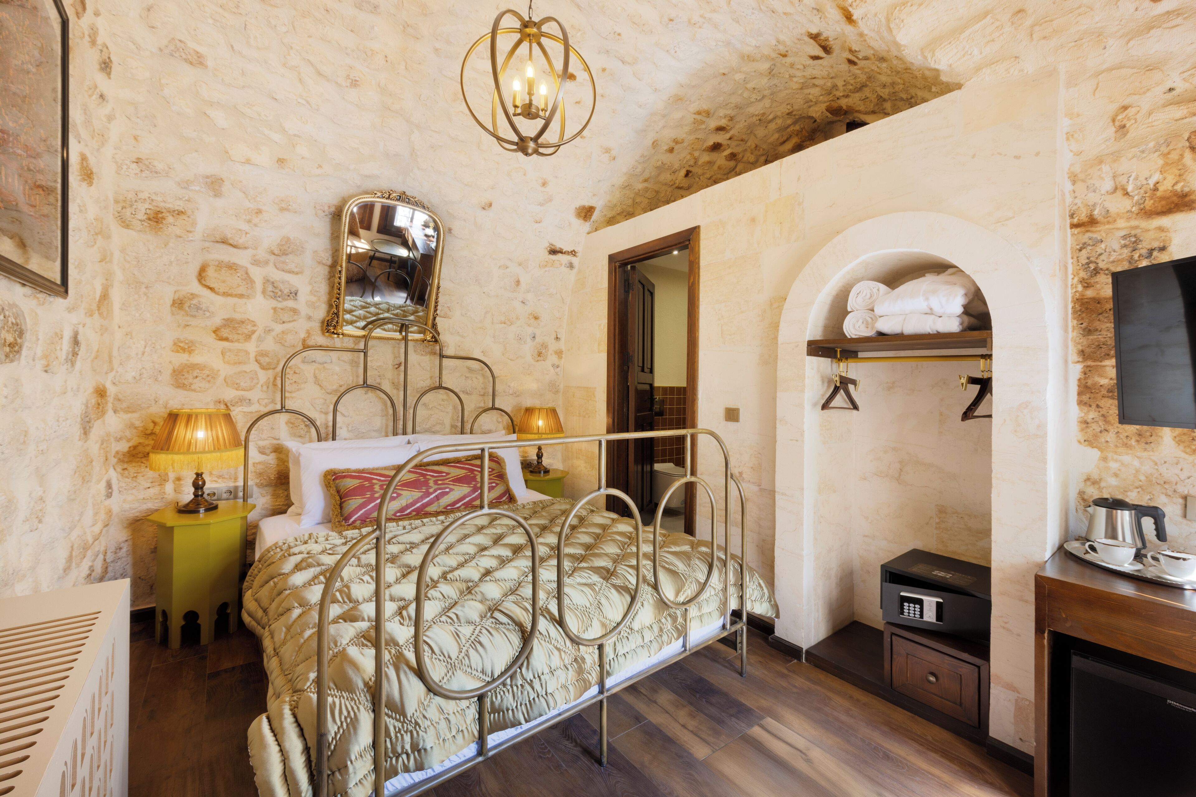 A bedroom with a stone wall, a bed with a metal frame, a bedside table with a lamp, a wooden cabinet, and a television on the wall.