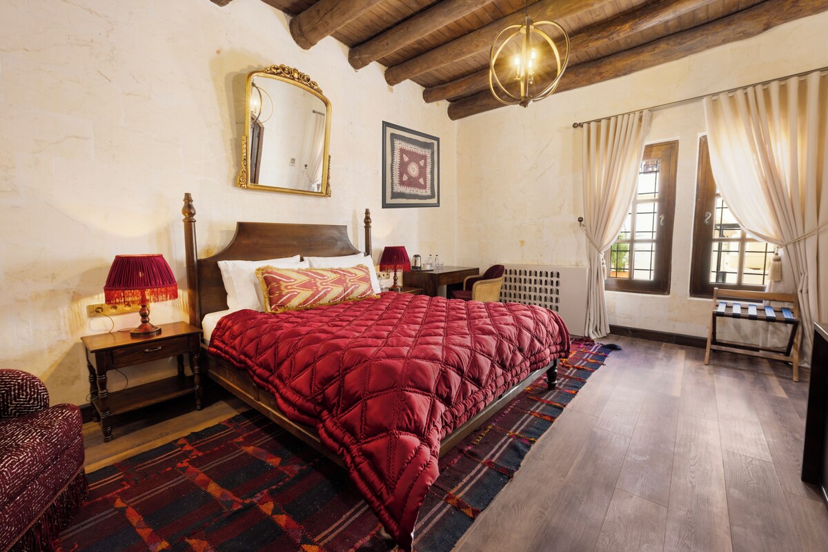 Un dormitorio con una cama grande, cabecera de madera, un espejo, una mesita de noche con lámpara, un radiador y una ventana con cortinas.