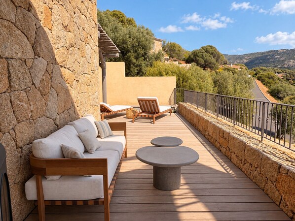 A deck with a sofa, chairs, and round tables, overlooking a scenic landscape with trees and hills.