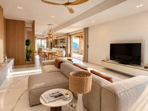 A modern living room with a sofa, a coffee table, a television, and a dining area with a kitchen in the background. - Villa Gemelli (Cargèse)
