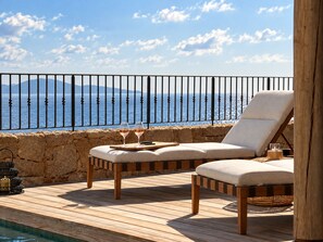 A wooden deck with two cushioned chairs, a table with a glass of wine, and a view of the sea.