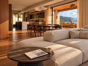 A modern living room with a large sectional sofa, a round coffee table with a newspaper, and a view of a dining area and mountains. - Villa Gemelli (Cargèse)