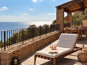 A wooden deck with a stone wall, a lounge chair, and a table set for dining with wine glasses. - Villa Gemelli (Cargèse)