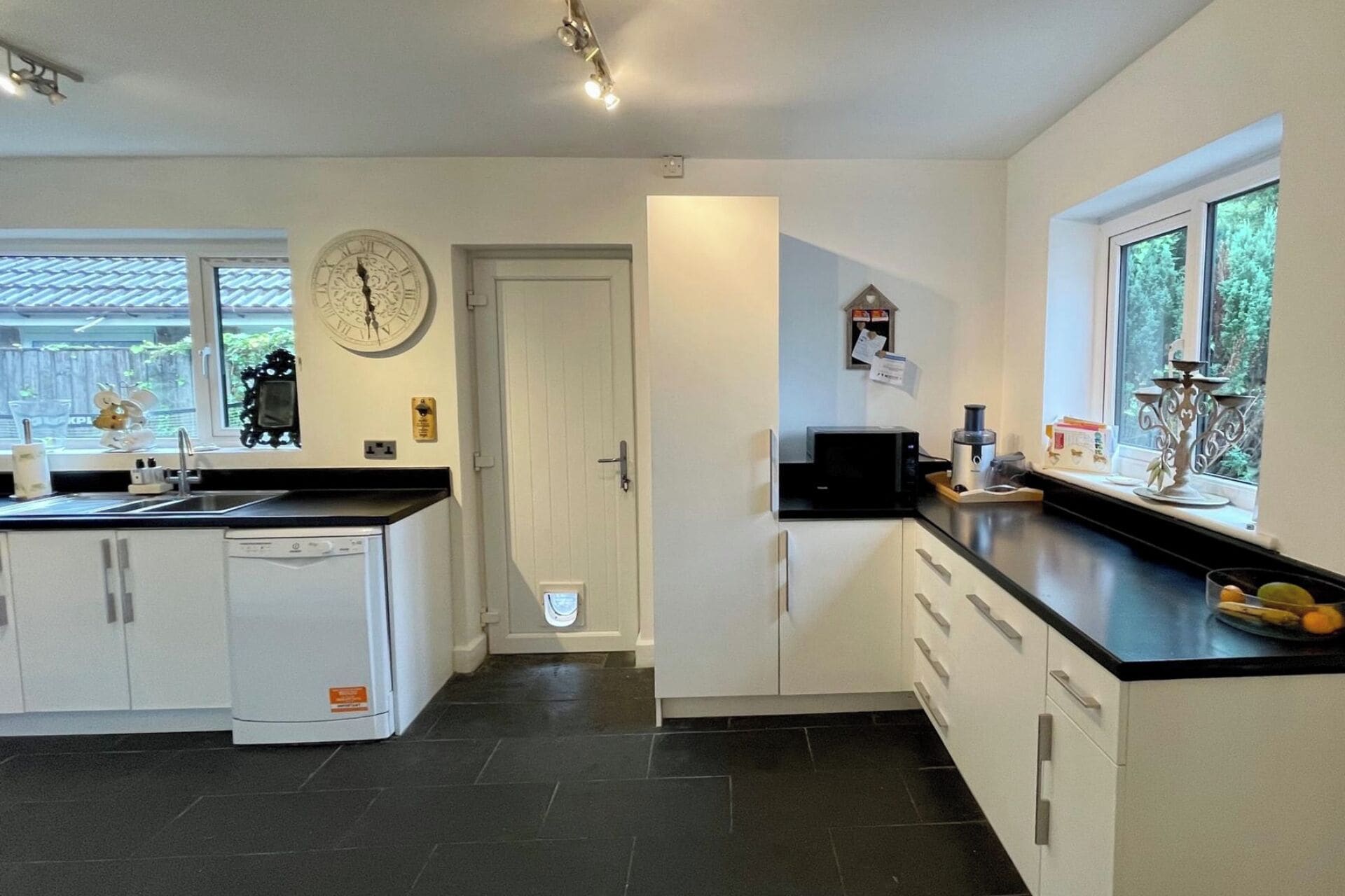 A modern kitchen with white cabinets, a dishwasher, a microwave, and a sink. There is a clock on the wall and a window with a view of greenery outside.