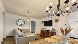 A living room with a sofa, a wooden dining table with placemats, a chandelier, a flat-screen TV, and a mirror on the wall. - 4 Bedroom House, Sleeps 8- Hot Tub, Pets, Parking (Altrincham)