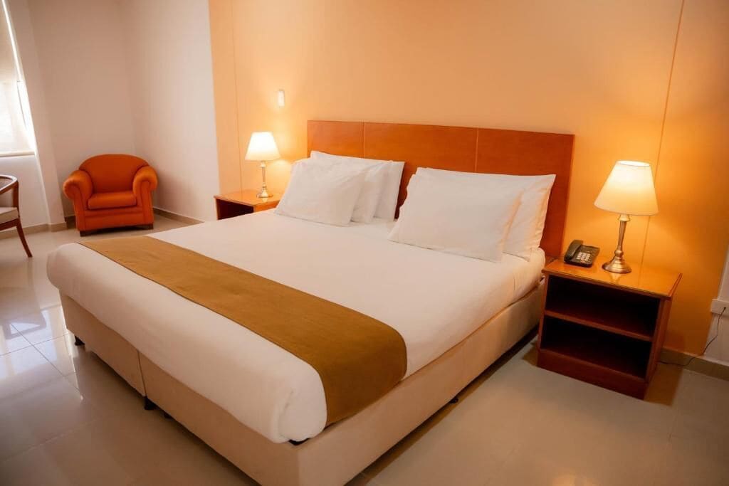 A hotel room with a large bed, two bedside tables with lamps, a chair, and a wooden headboard.