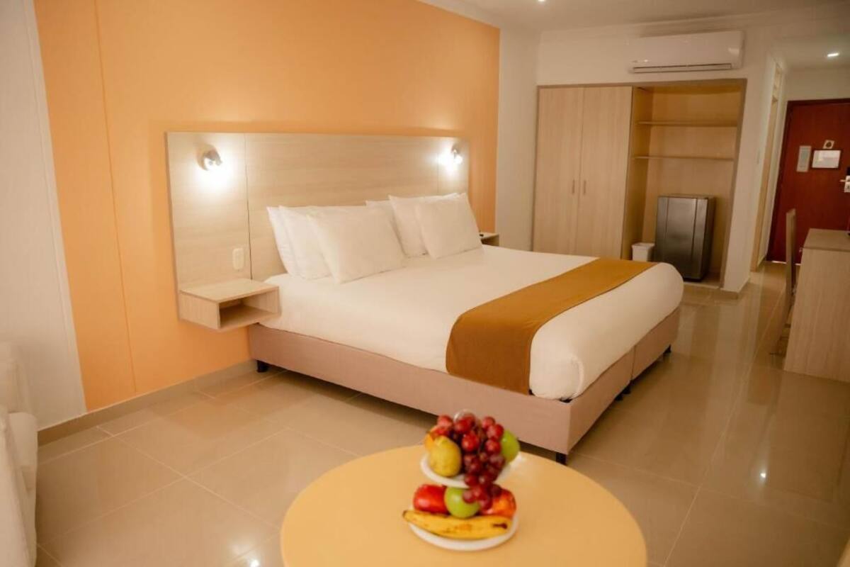 A modern hotel room with a large bed, a bedside table, a round table with fruit, and a wardrobe.