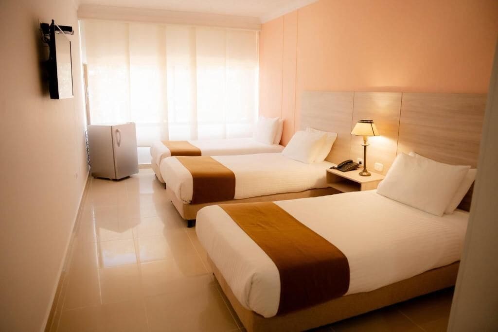 Habitación de hotel con dos camas, una mesita de noche con lámpara, un refrigerador y una ventana con persianas.