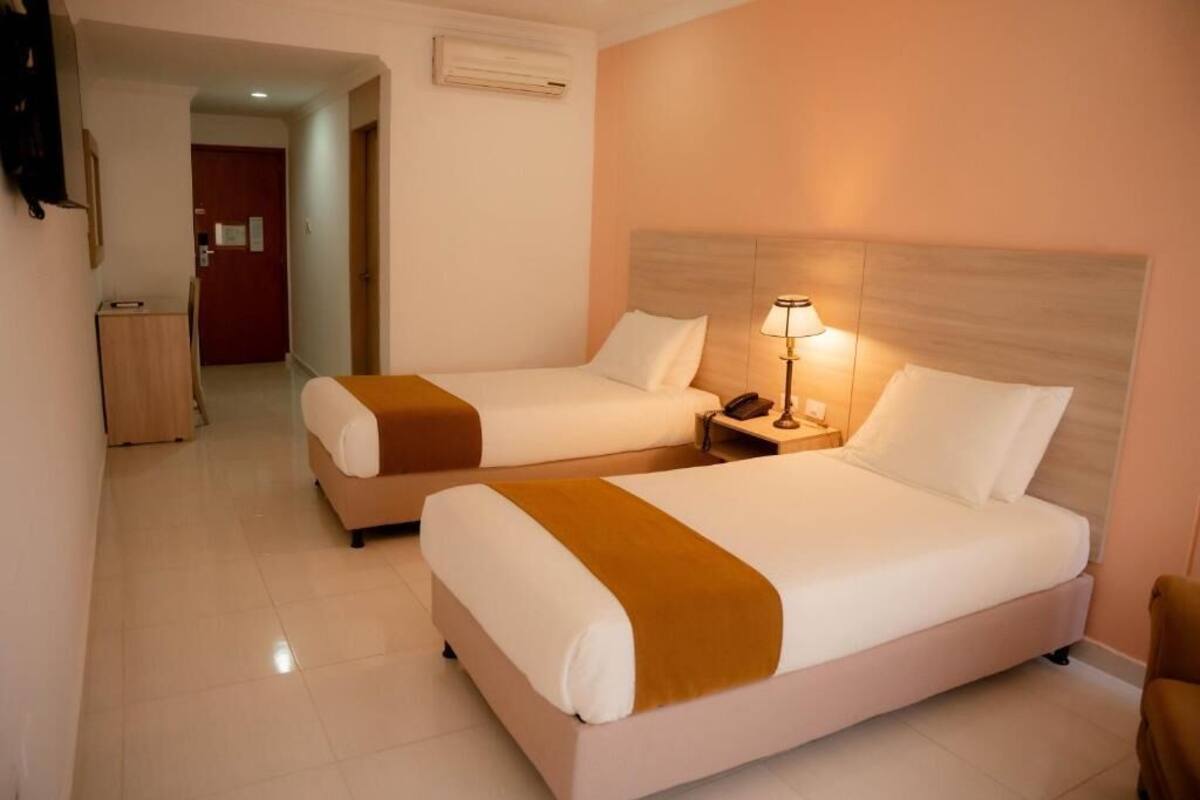 A hotel room with two beds, a nightstand with a lamp, and a door leading to a hallway.