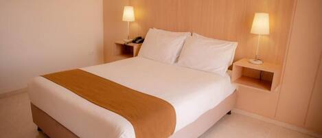 Una cama bien tendida con sábanas blancas y una manta marrón, con dos mesitas de noche a cada lado, cada una con una lámpara, en una habitación con cabecera de madera.