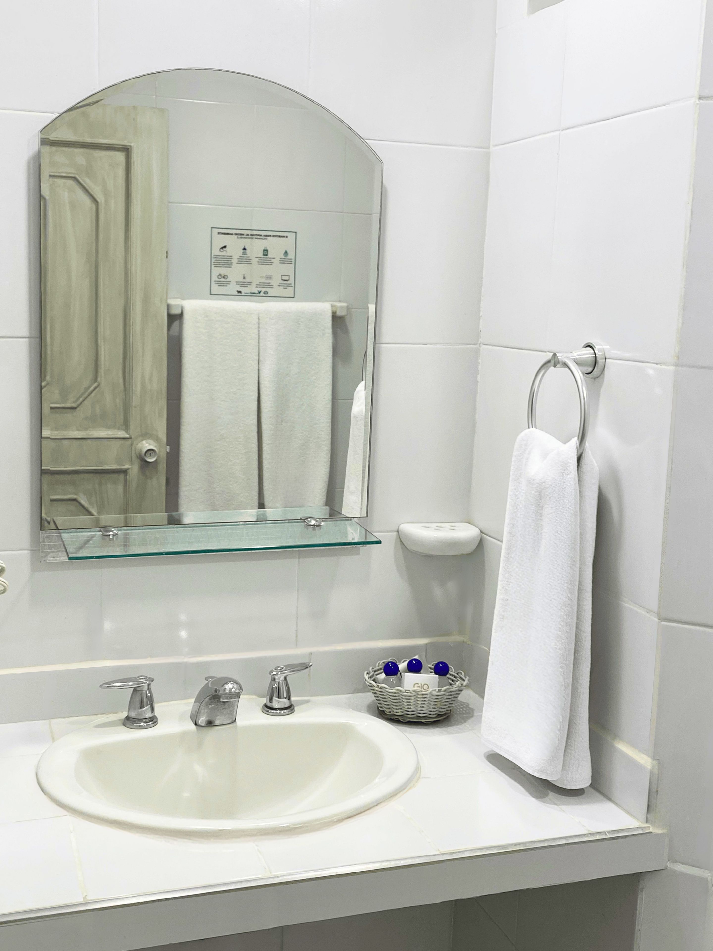 A bathroom with a white sink, two faucets, a glass shelf above the sink, a towel rack, and a mirror.