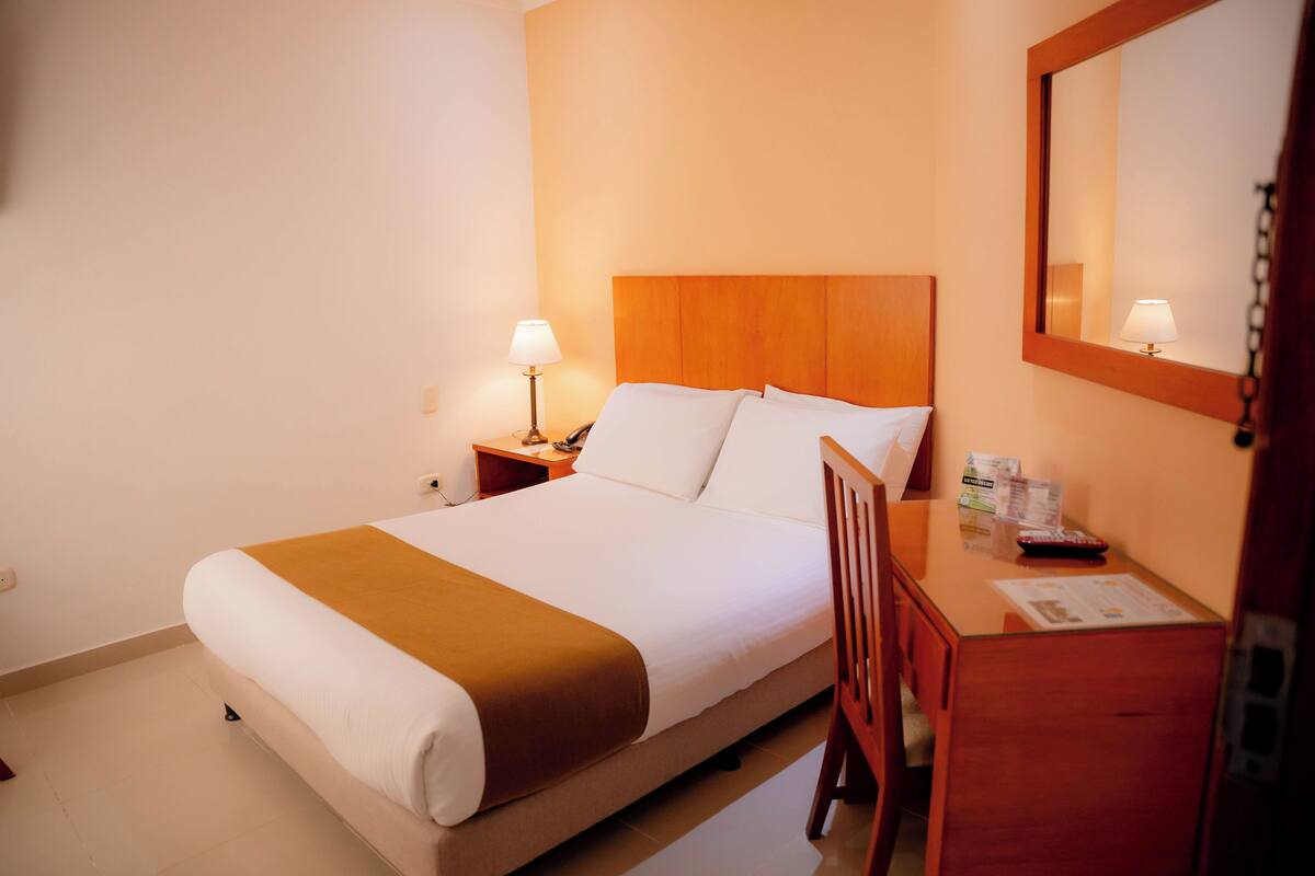 A neatly made bed with white and brown linens, a wooden headboard, a bedside table with a lamp, and a mirror on the wall.