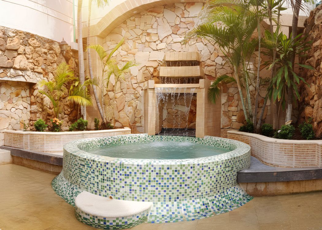 A hot tub with a mosaic tile design, surrounded by a stone wall and palm trees.