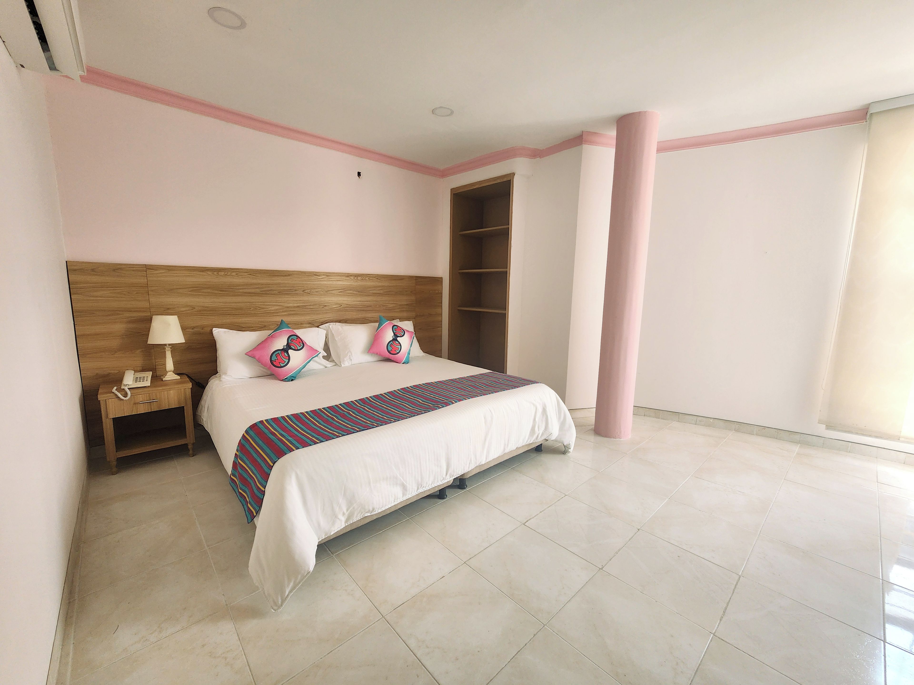 Una habitación de hotel con una cama grande, una mesita de noche con lámpara y teléfono, un cabecero de madera y un ventanal amplio.