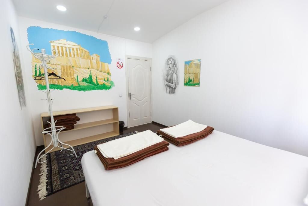 A hotel room with a bed, a bench, a coat rack, and a wall painting of a historical building.