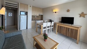 Un salon moderne comprenant un canapé, une table basse avec une plante, un coin repas avec des chaises, une cuisine équipée et un téléviseur posé sur un meuble en bois.