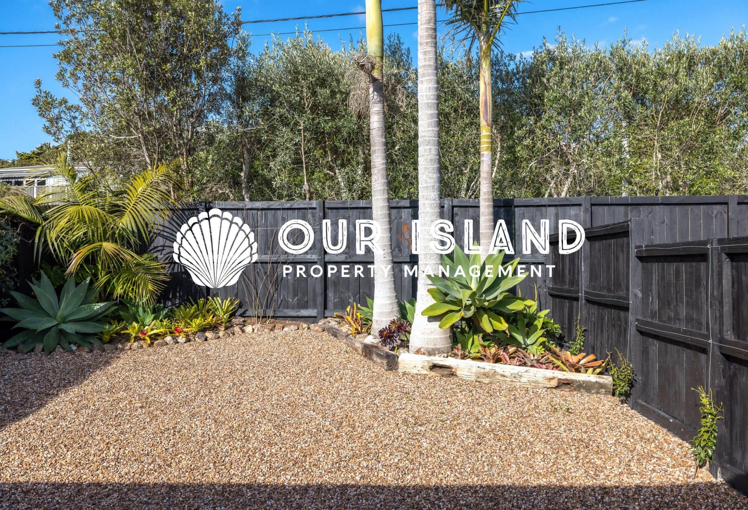 A property management sign with a shell logo and the text "OUR ISLAND PROPERTY MANAGEMENT" in front of a wooden fence with tropical plants.