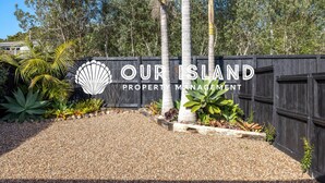 Skilt fra et ejendomsforvaltningsfirma med et logo af en snegl og teksten "OUR ISLAND PROPERTY MANAGEMENT" foran et træhegn med tropiske planter.