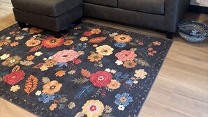 A living room with a dark-colored sectional sofa, a floral-patterned rug, and a painting on the wall.