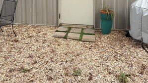 A metal building with a door labeled "The Goat Shed," a small step leading up to the door, a chair, and a potted plant.