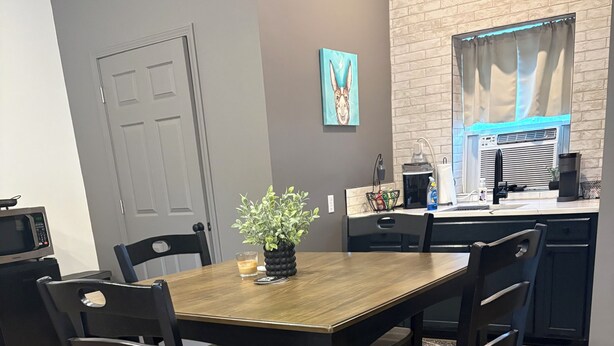 A dining area with a wooden table and chairs, a potted plant, and a kitchen in the background.