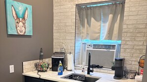 A kitchen with a white countertop, black cabinets, a window with curtains, a wall-mounted air conditioner, and a painting of a rabbit.