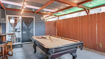 Una mesa de billar con estructura de madera y tela de color claro, rodeada por una valla de madera y una estructura de techo metálica.