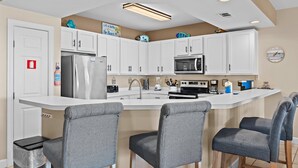 A modern kitchen with white cabinets, a stainless steel refrigerator, a microwave, and a sink. There are three grey bar stools and a trash can. - Pavilion Palms 105c- The Emerald Jewel (Miramar Beach)