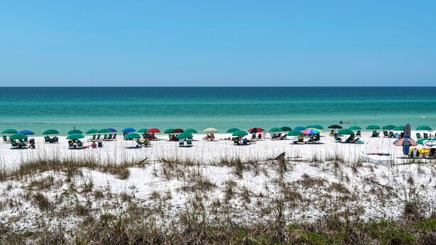 A beach with rows of beach chairs and umbrellas, a sandy foreground with grass, and the ocean in the background. - Pavilion Palms 105c- The Emerald Jewel (Miramar Beach)