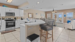 A modern kitchen with white cabinets, a microwave, and a stove. There is a dining area with a white table and chairs. - Pavilion Palms 105c- The Emerald Jewel (Miramar Beach)
