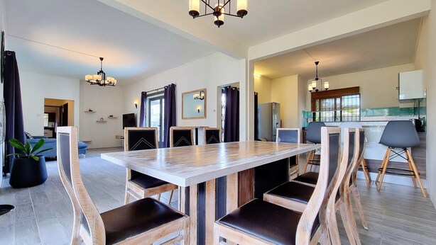 A spacious dining area with a large wooden table and chairs, a modern kitchen in the background, and a view of the living area. - Royal Residence A3 - Gated Semi Detached Spacious Residence in Pereybere (Grand Baie)