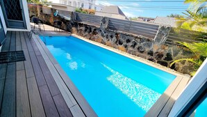 A swimming pool with a wooden deck, stone walls, and a view of buildings in the background. - Royal Residence A3 - Gated Semi Detached Spacious Residence in Pereybere (Grand Baie)