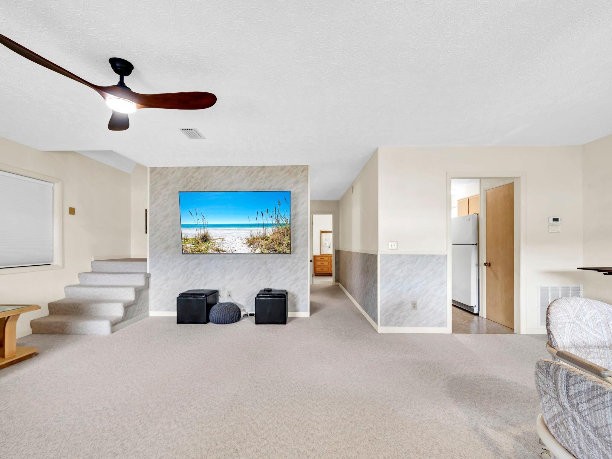A modern living room with a large flat-screen TV mounted on a wall, a ceiling fan, a staircase leading to another level, and a view of a beach through the TV.