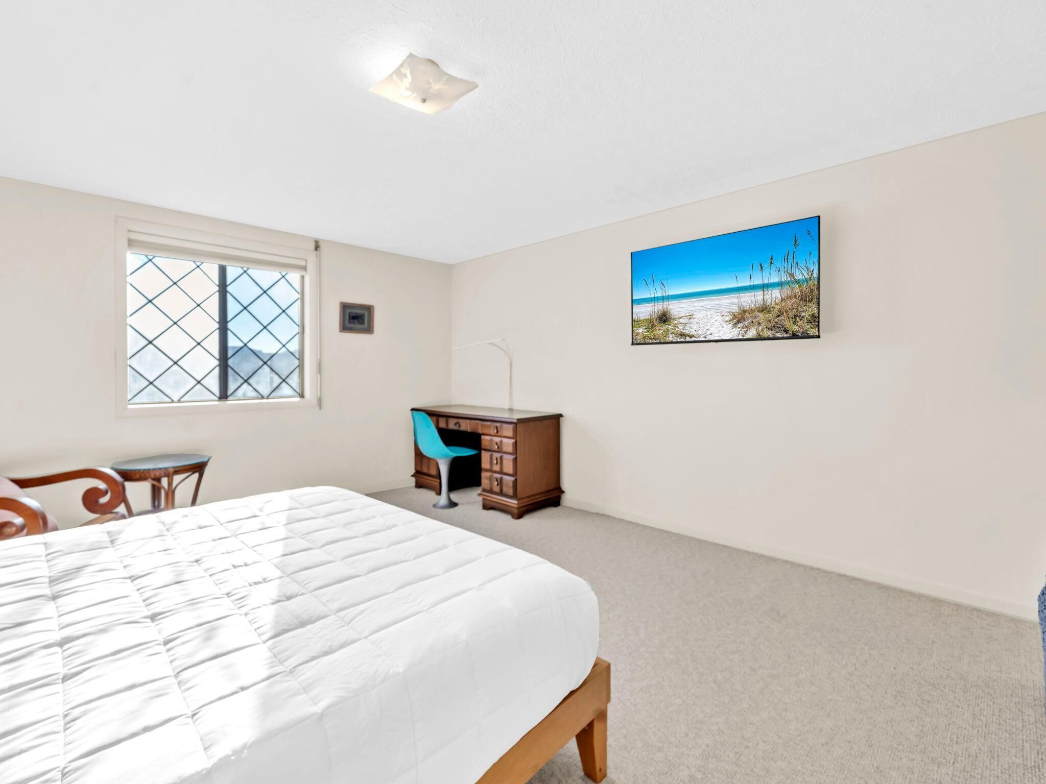 A bedroom with a bed, a desk, a chair, a window, and a beach scene painting.