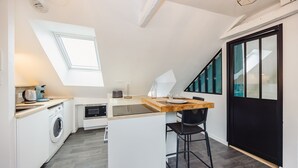 A compact kitchen with a built-in washing machine, a small dining area, and a black-framed sliding door. - Le Cosy Comble - 10 min Campus Kerlann (Bruz)