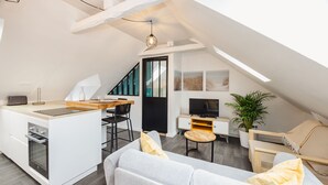 A modern loft apartment with a kitchen, dining area, and living room. It features a large window, a staircase, and a flat-screen TV. - Le Cosy Comble - 10 min Campus Kerlann (Bruz)