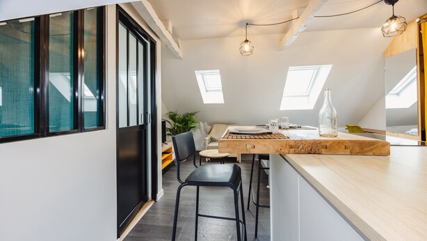 A modern kitchen with a wooden countertop, black stools, and a dining table. - Le Cosy Comble - 10 min Campus Kerlann (Bruz)