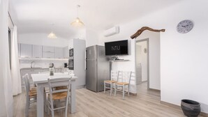 A modern kitchen with white cabinets, stainless steel appliances, a dining table with chairs, and a TV mounted on the wall. - Independent Domu Fama (Tertenia)