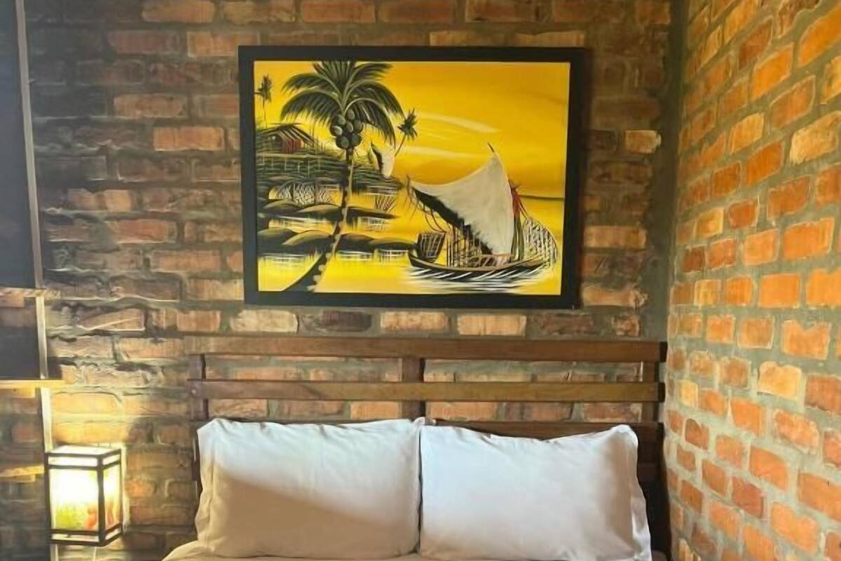 A room with a brick wall, a bed with white and red bedding, and a painting of a tropical scene.
