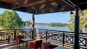 A covered outdoor seating area with tables and chairs, overlooking a lake with boats and trees. - Hotel Niedzwiadek (Koscierzyna)