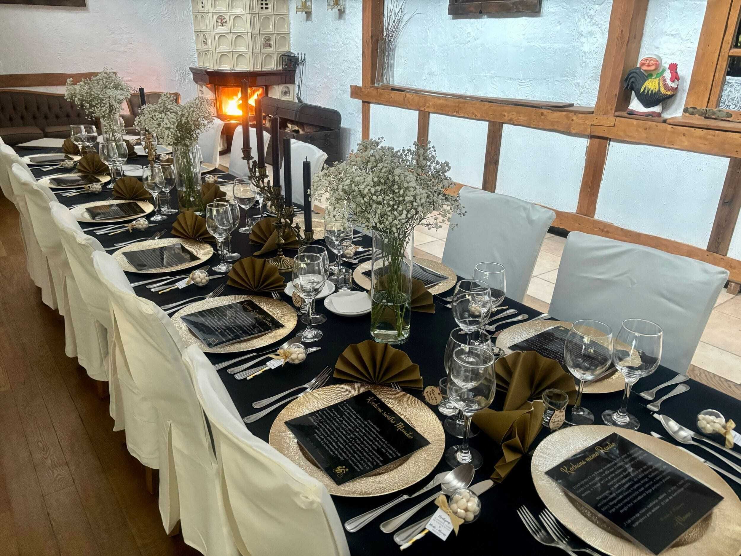 A long dining table set for a formal event with gold and black tableware, white tablecloths, and a fireplace in the background.