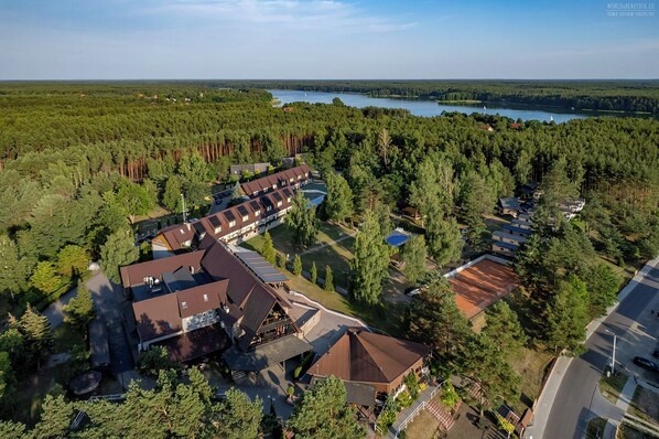 Vue aérienne d’un complexe hôtelier comprenant plusieurs bâtiments, un court de tennis et un lac en arrière-plan.