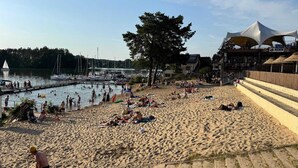 A beach with people sunbathing and swimming, a dock with boats, and a building with a white canopy. - Hotel Niedzwiadek (Koscierzyna)