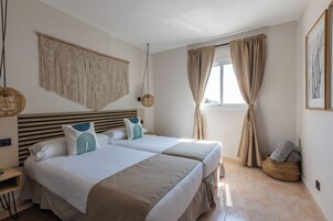 Un dormitorio con dos camas, un colgador de macramé, una lámpara tejida y una ventana con cortinas.