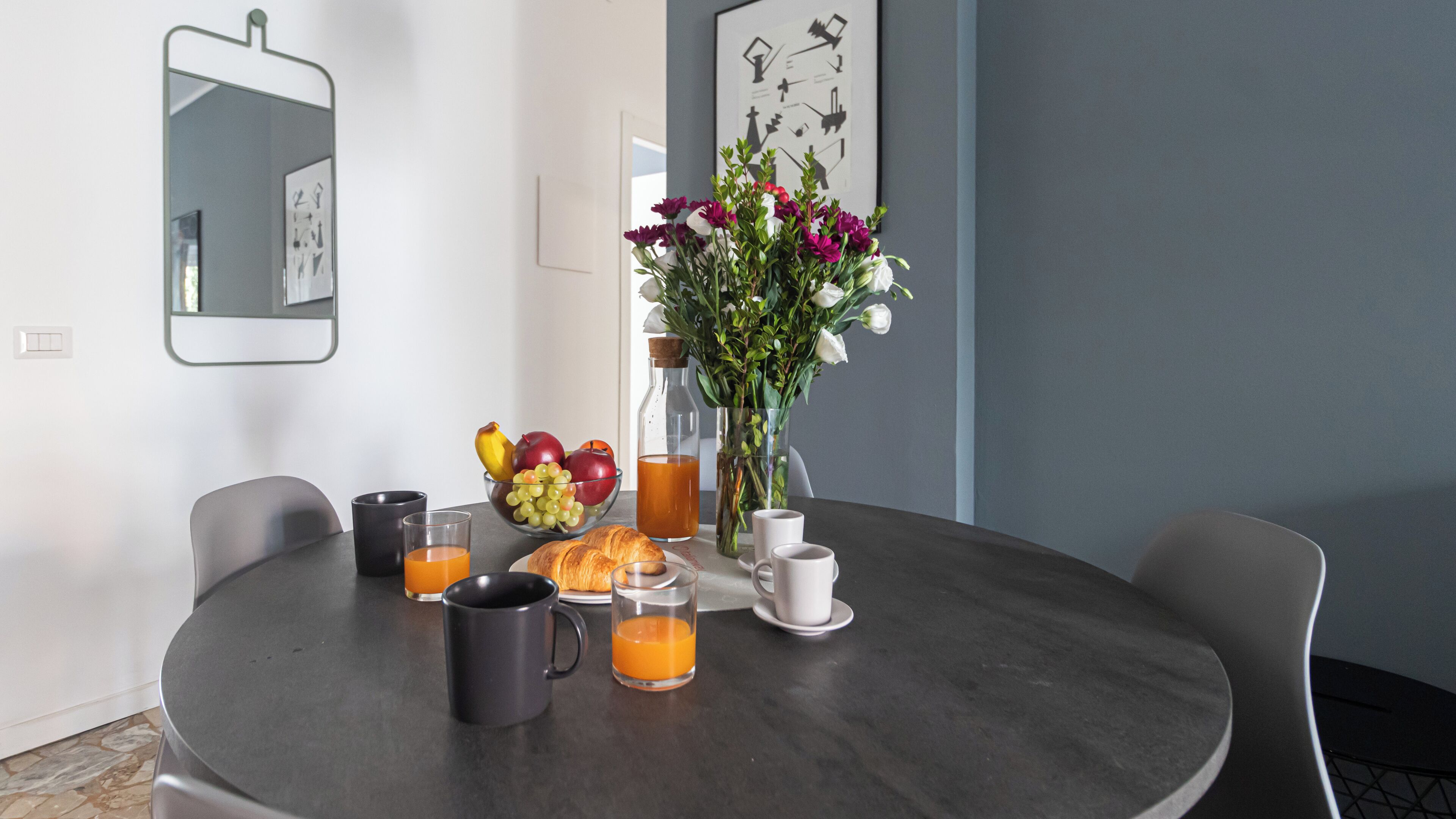 A round dining table with a breakfast setting including croissants, fruit, juice, and a vase with flowers.