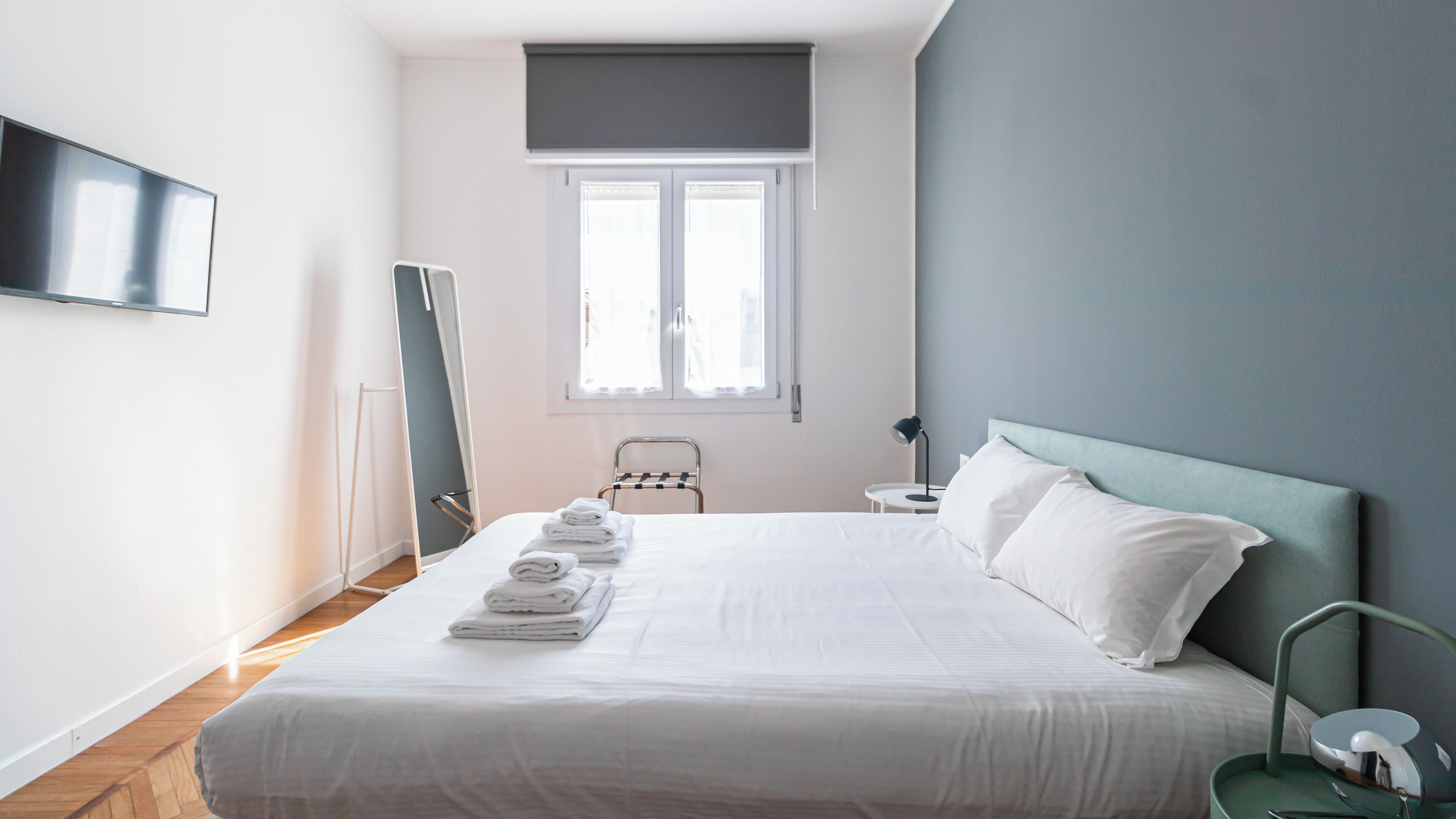 Een slaapkamer met een bed, een aan de muur bevestigde televisie, een raam met jaloezieën, een nachtkastje met een lamp en een stoel.